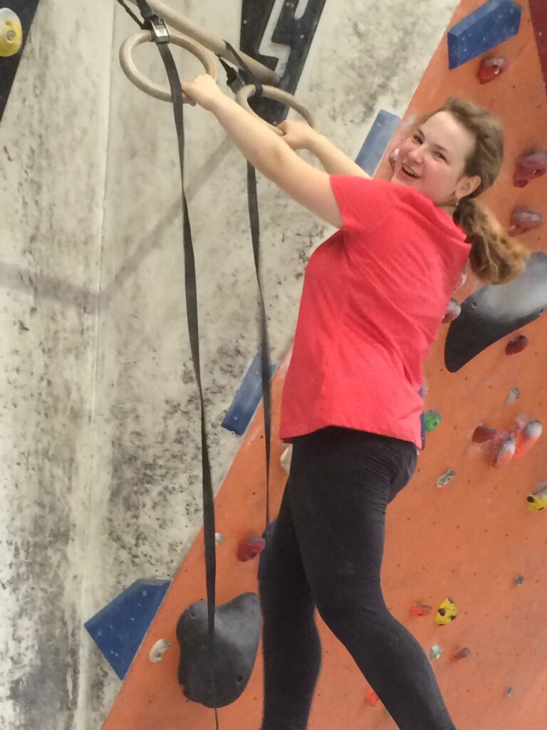 Larissa Lang beim Bouldern / Klettern, sie hält sich gerade an Ringen fest und lächelt zufrieden in die Kamera