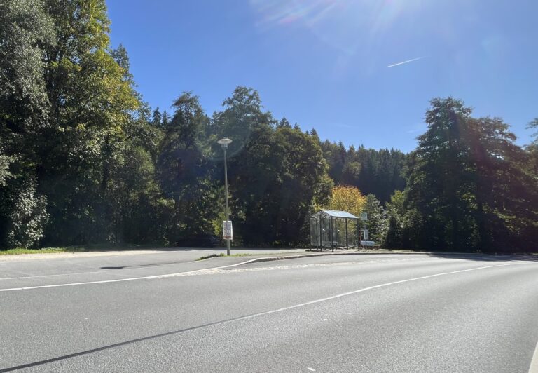 Bild eines geschotterten Parkplatzes hinter einer Bushaltestelle mit Bushäuschen und Mitfahrbank, von der gegenüberliegenden Straßenseite aus fotografiert