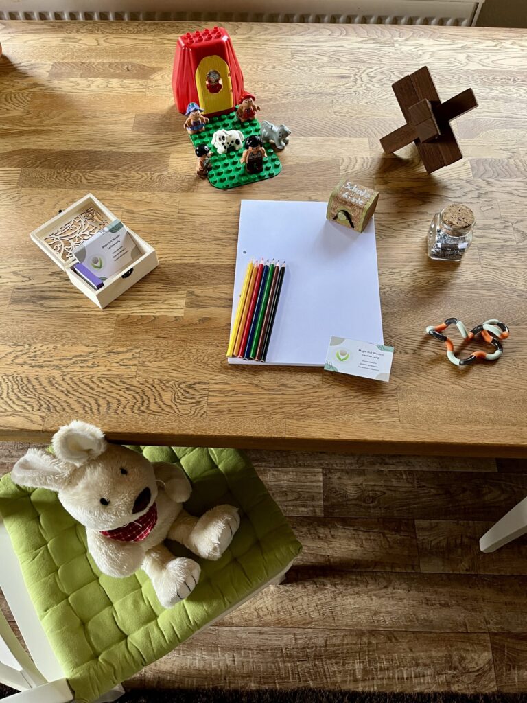 Friedolin die Hasenhandpuppe sitzt auf einem grünen Sitzkissen, welches auf einem weißen Stuhl liegt, dabei lehnt er mit dem Kopf an dem Holztisch, auf welchem zum einen Buntstifte, ein weißer Block, eine kleine Schatzkiste, ein Knobelspiel, ein Fidgettoy, eine kleine Legoplatte mit Figuren und Tieren, sowie einer Tür liegen. Außerdem ist dort noch eine Kiste mit Visitenkarten und kleinen lilanen Notizzettelchen.