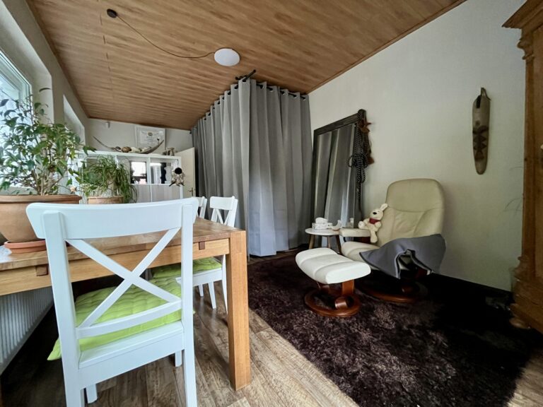 Foto von der Praxis: Auf dem Foto sind der beige farbene Stressless Sessel mit dem dazugehörigen Fußteil, auf einem der Armlehnen liegt eine graue Decke und auf der anderen Lehne sitzt die Hasenhandpuppe Friedolin, ein Beistelltisch mit einer weißen Salzkristalllampe in Mondform und einer Taschentücherbox, ein brauner Teppich, Spiegel an dem ein Halfter eines Pferdes hängt, ein großer Holzschrank, weiße Stühle und ein Holztisch, auf dem eine Pflanze steht, sowie ein Regal, graue Vorhänge und ein Zertifikat über der Tür zu sehen.