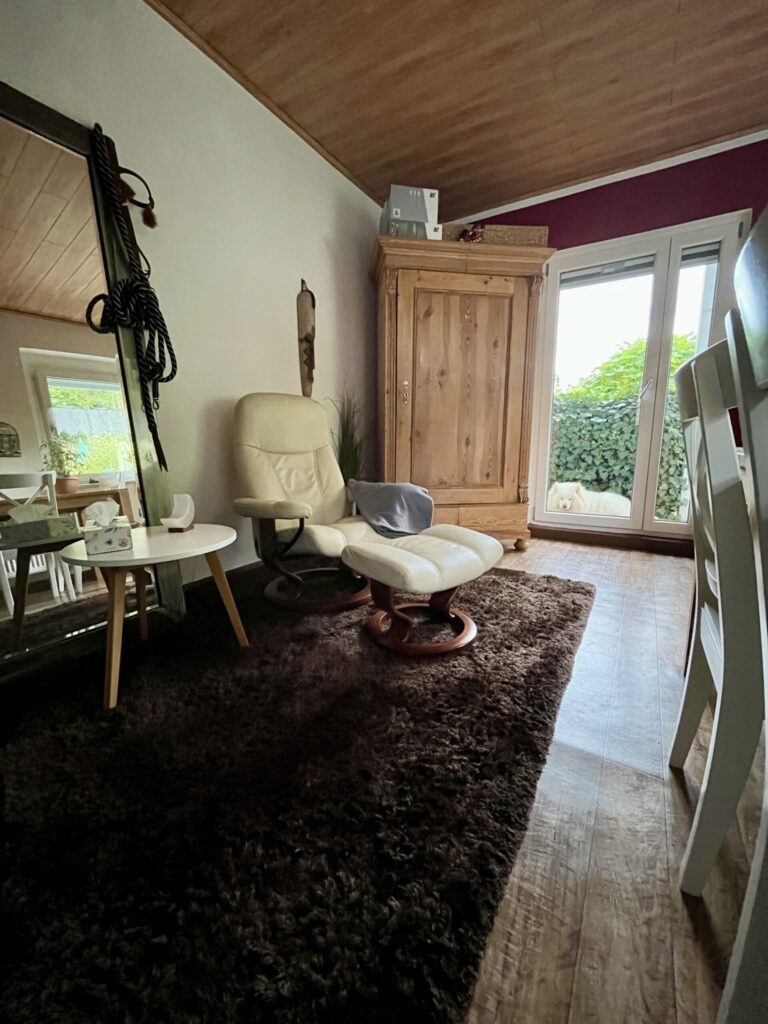 Foto von der Praxis: Auf dem Foto sind der beige farbene Stressless Sessel mit dem dazugehörigen Fußteil, auf einer der lehnen liegt eine graue Decke, ein Beistelltisch mit einer weißen Salzkristalllampe in Mondform und einer Taschentücherbox, ein brauner Teppich, Spiegel an dem ein Halfter eines Pferdes hängt, ein großer Holzschrank, weiße Stühle und die weiße Praxishündin Sari (Samoyede) zu sehen.