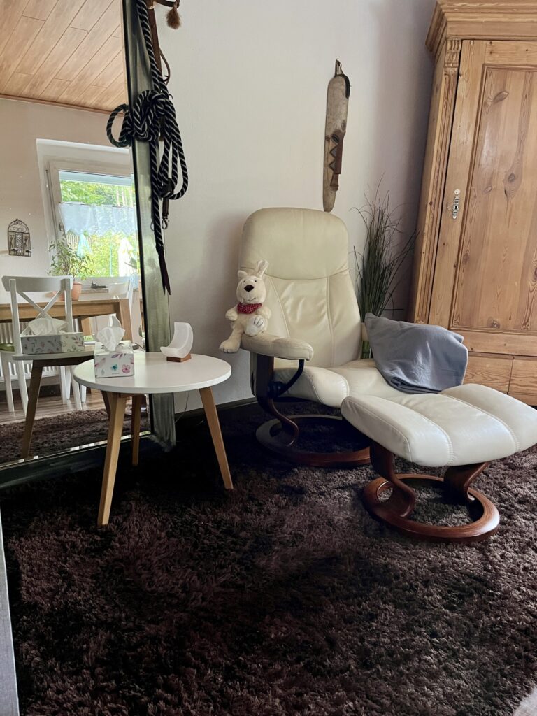 Foto von der Praxis: Auf dem Foto sind der beige farbene Stressless Sessel mit dem dazugehörigen Fußteil, auf einer der Lehnen des Sessels, liegt eine graue Decke und auf der anderen Lehne sitzt die Hasenhandpuppe Friedolin, ein Beistelltisch mit einer weißen Salzkristalllampe in Mondform und einer Taschentücherbox, ein brauner Teppich, Spiegel an dem ein Halfter eines Pferdes hängt und ein großer Holzschrank zu sehen.